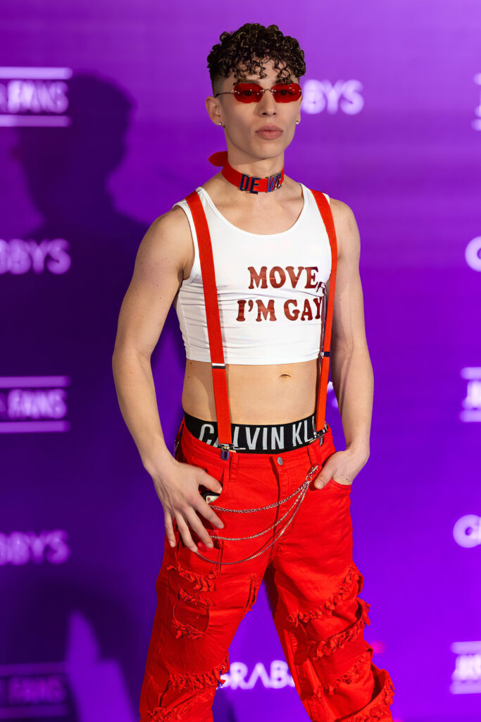 Creator David de Vil posing in a red-themed outfit with crop top, suspenders and red sunglasses on the Purple Carpet at Grabbys Europe 2026.