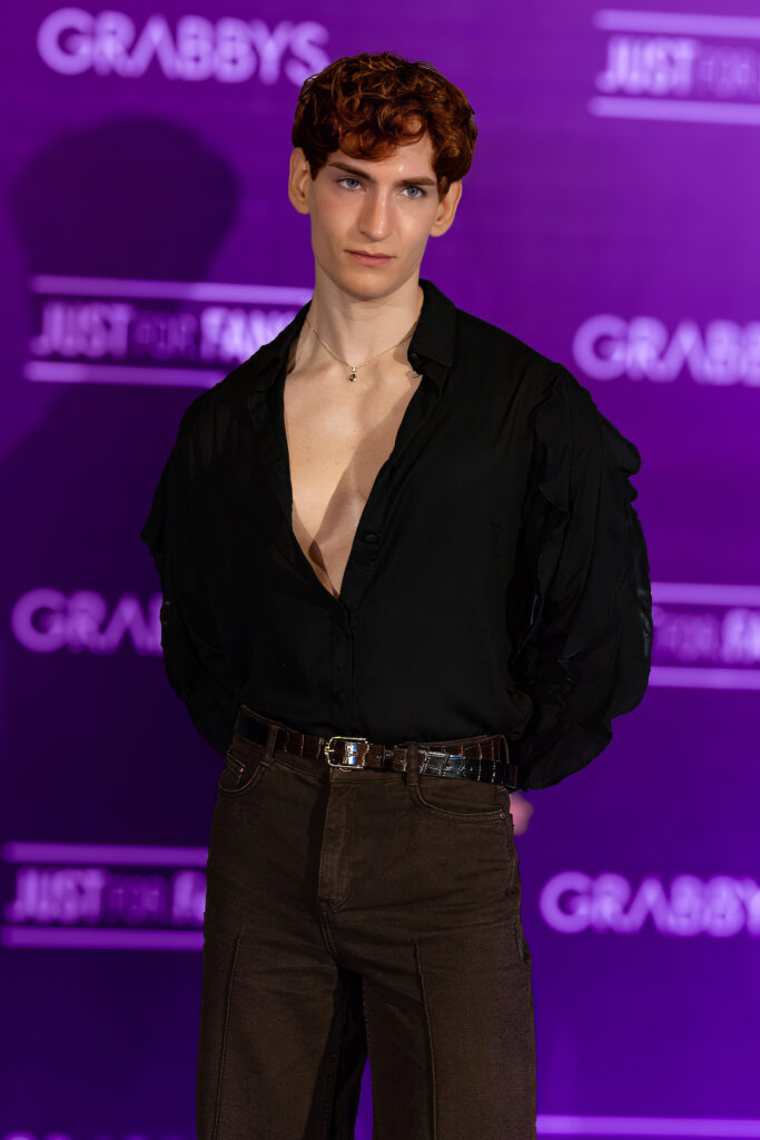 British creator Evander Johnson posing in a black shirt with a deep neckline and brown trousers on the Purple Carpet at Grabbys Europe 2026.