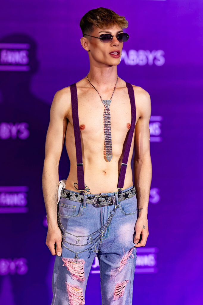 British creator Roby de Vil posing shirtless with glittering tie, red suspenders and distressed jeans on the Purple Carpet at Grabbys Europe 2026.